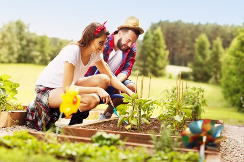 A man and a woman are working together in a well-maintained garden bed in the outdoor space of a residential property, with a lush, green landscape featuring trees and grass in the background. The garden bed contains various young plants and sprouts, covered with rich, dark soil and bordered by wooden planks. The woman, wearing a white t-shirt and plaid shorts, is smiling and tending to the plants, while the man, dressed in a red and blue checkered shirt and straw hat, assists nearby. Bright, natural daylight highlights the vibrant shades of green and earthy tones, emphasizing a peaceful gardening scene. The surrounding area includes a gravel pathway and a glimpse of a garden ornament in the form of a small, colorful bird figurine, enhancing the aesthetic appeal and connection to outdoor gardening services such as planting and garden maintenance. The environment appears warm and sunny, ideal for outdoor activities, reflecting the natural and sustainable approach typical of gardening specialists serving Newham and surrounding areas.