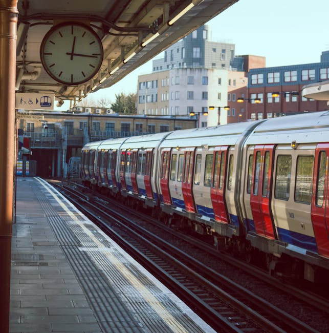 A railway station platform with a weathered metal canopy supported by teal-painted pillars showing peeling paint. The platform surface is paved with grey tiles, featuring tactile paving strips for vis